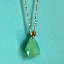 Charger l&#39;image dans la galerie, Gros plan sur sautoir talisman bohème avec perle en verre opaque vert tendre taillé irrégulier et galet doré à l&#39;or fin sur fil doré. Fond bleu turquoise.
